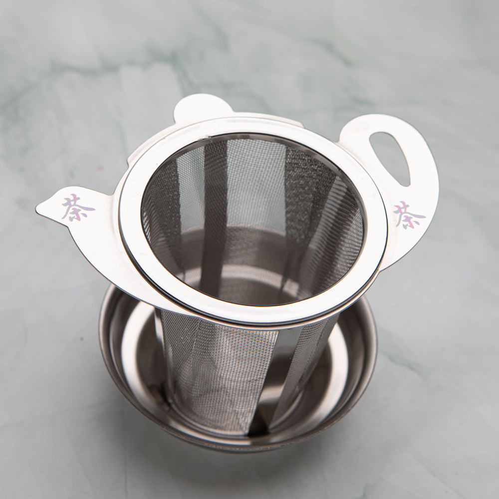 Stainless steel tea strainer with a teapot shaped lid marked with the character for tea, on a drip dish, by Bettina's Tea