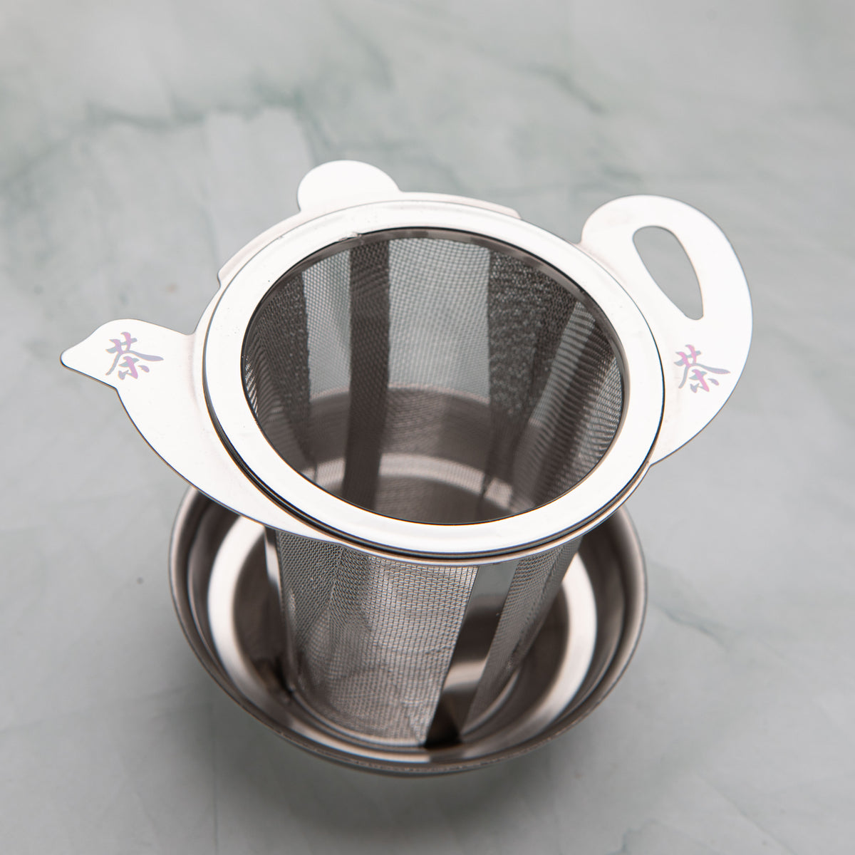 Stainless steel tea strainer with a teapot shaped lid marked with the character for tea, on a drip dish, by Bettina's Tea