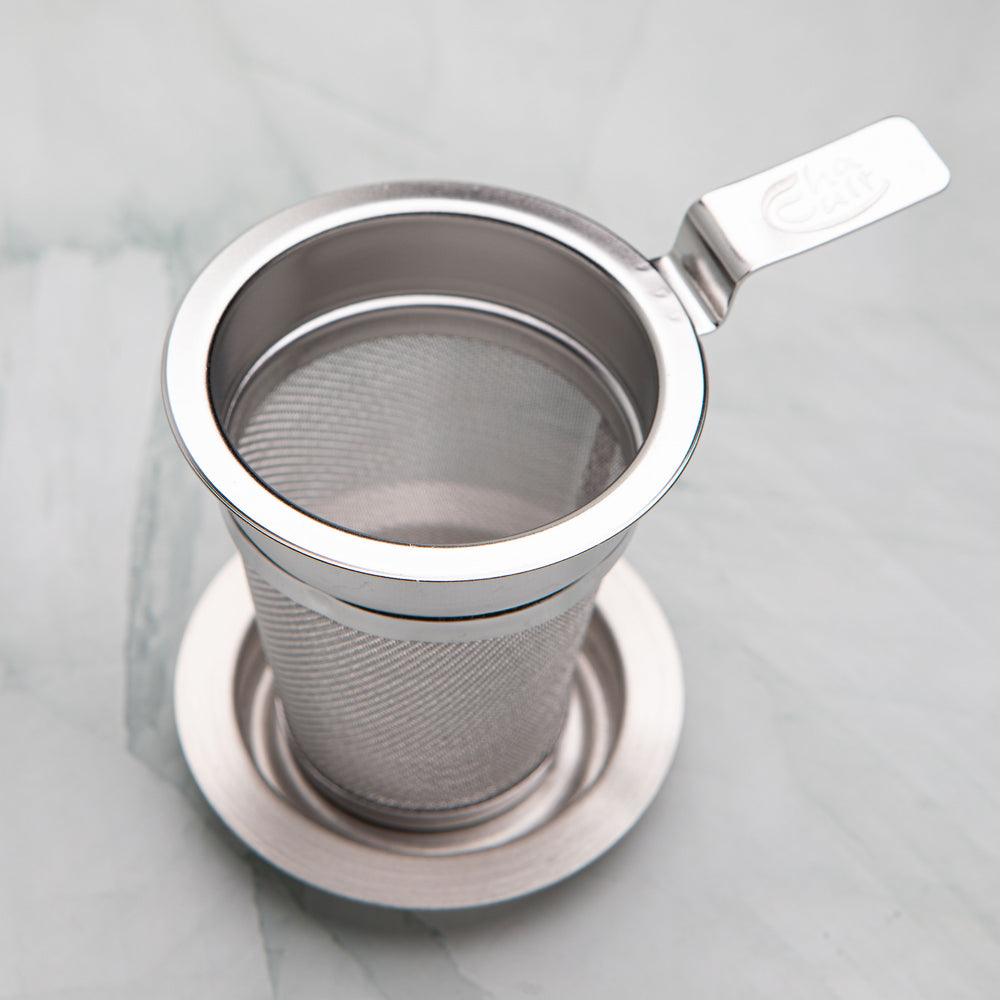 Stainless steel tea strainer with a flat handle resting on its dish, by Bettina's Tea