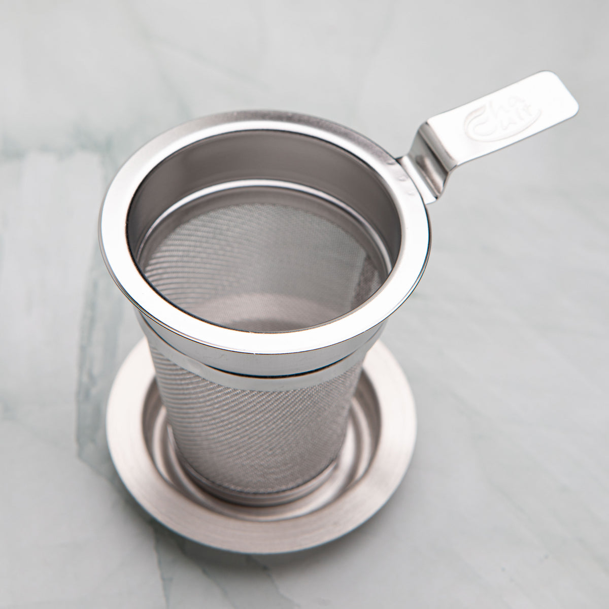 Stainless steel tea strainer with a flat handle resting on its dish, by Bettina's Tea