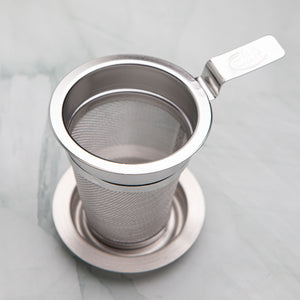 Stainless steel tea strainer with a flat handle resting on its dish, by Bettina's Tea