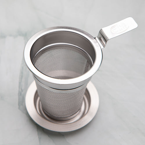 Stainless steel tea strainer with a flat handle resting on its dish, by Bettina's Tea
